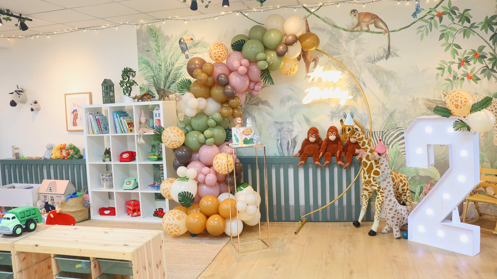 A balloon arch and Happy Birthday neon sign in front of Dreami's jungle mural